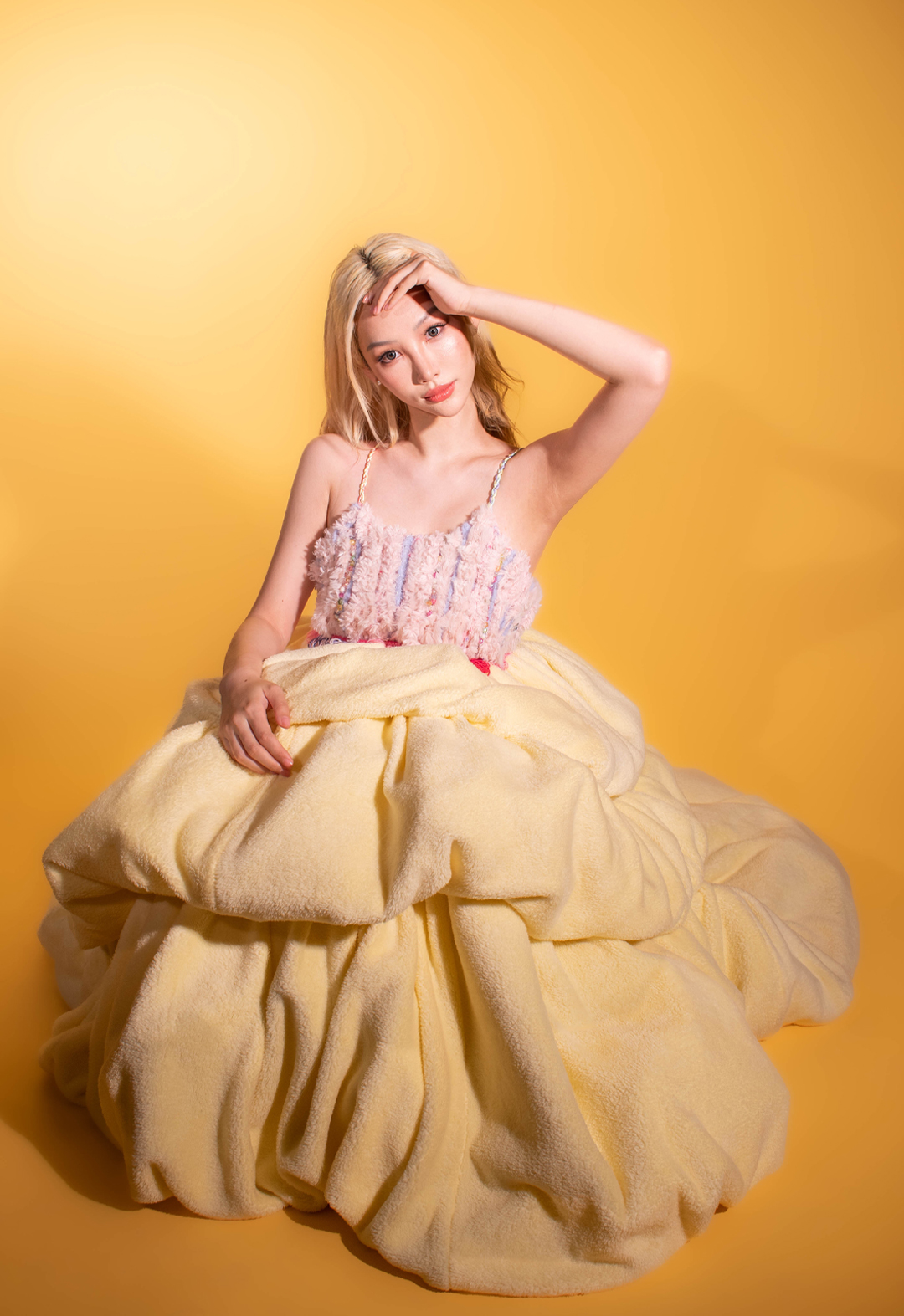 Photo of a girl with blonde hair sitting and facing to the camera. She has one of her hands on her head and another hand on the dress. She is wearing a sleeveless pastel-pink-and-yellow dress. It also has a bright-pink knit belt. The background of this picture is yellow.