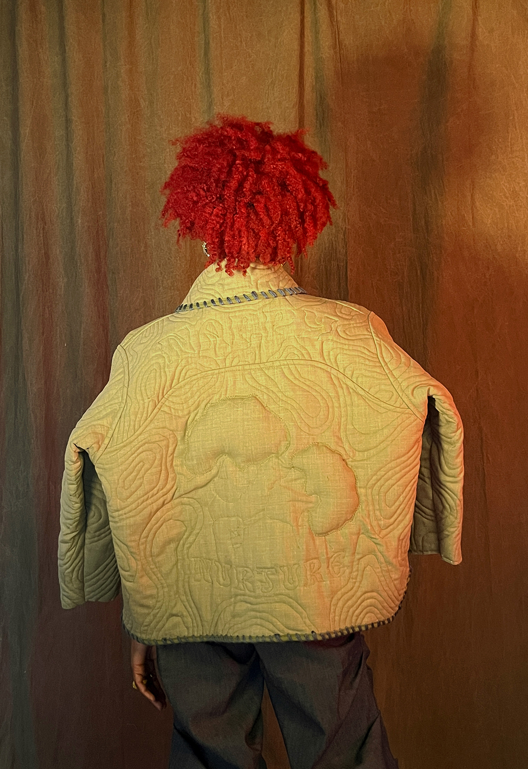 Back view of a model wearing a quilted jacket that shows the image of a mother and a young daughter with the words "Nappy" and "Nurture" surrounding them.
