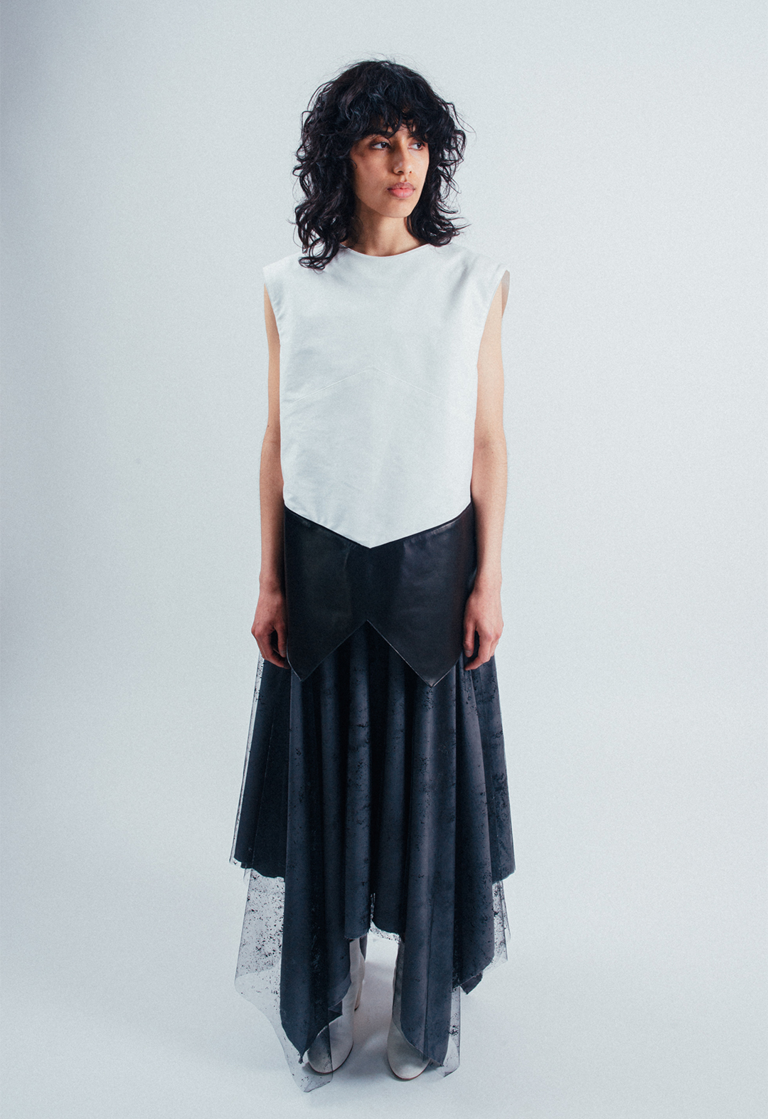 Photo of the model, Aria Puga, wearing a white cotton and black leather open-back top and a grey Tencel handkerchief skirt with black nylon tulle painted with enamel paint. The model is looking to the side. The background is white.