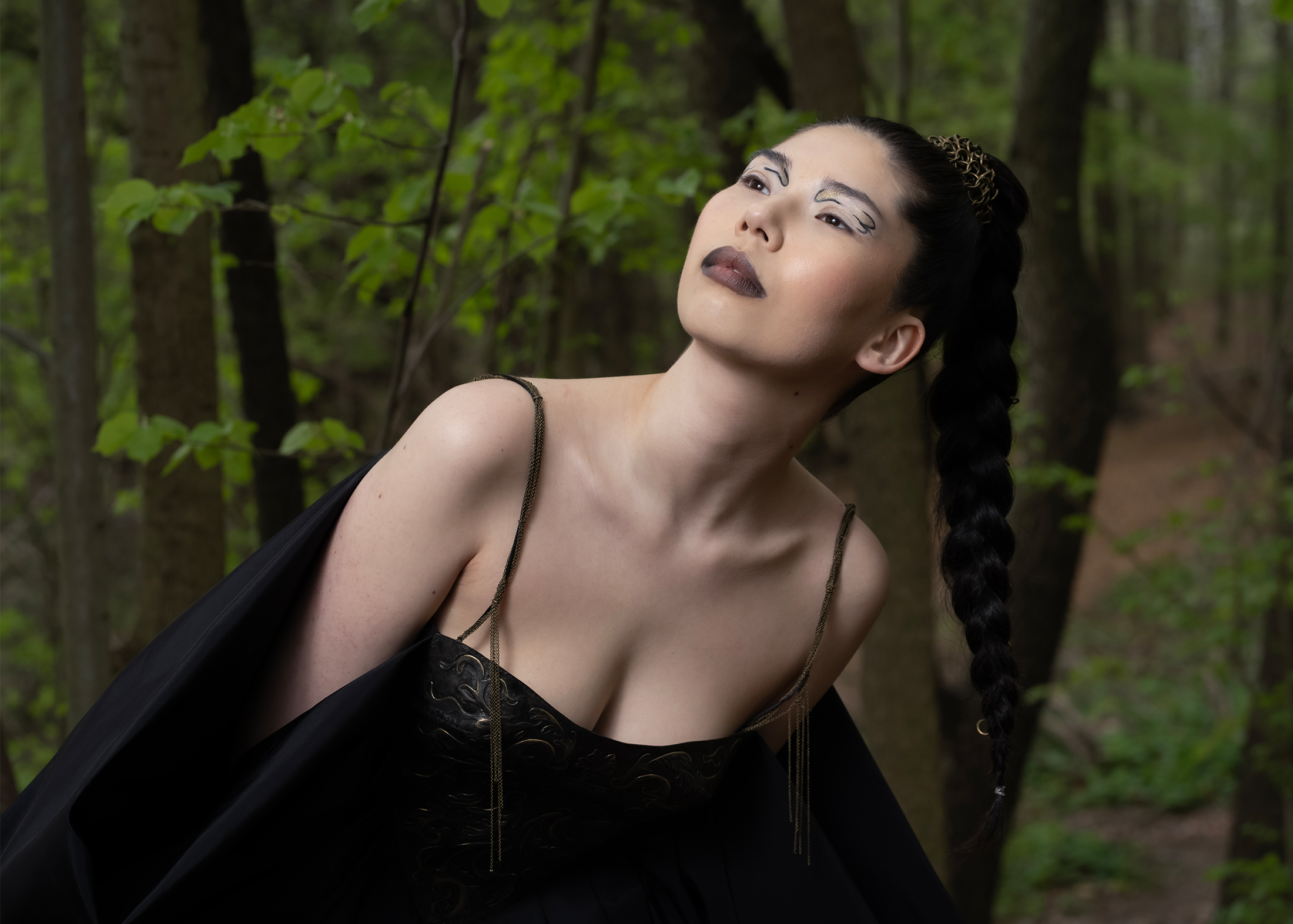 A woman in a black-and-gold dress in the forest is the focal point of this image, exuding an air of mystery and allure. The camera zooms in on various aspects of her appearance, capturing her intricate braid. Her makeup is dark and dramatic, adding to the enigmatic aura that surrounds her. The overall effect is one of sophistication and elegance, while capturing the details of the delicate hand-painted/leather-tooling bodice with its delicate chains as straps. The overall ambiance gives a strong woman/warrior vibe, one of a women exuding an air of quiet confidence, resilience, and bravery.