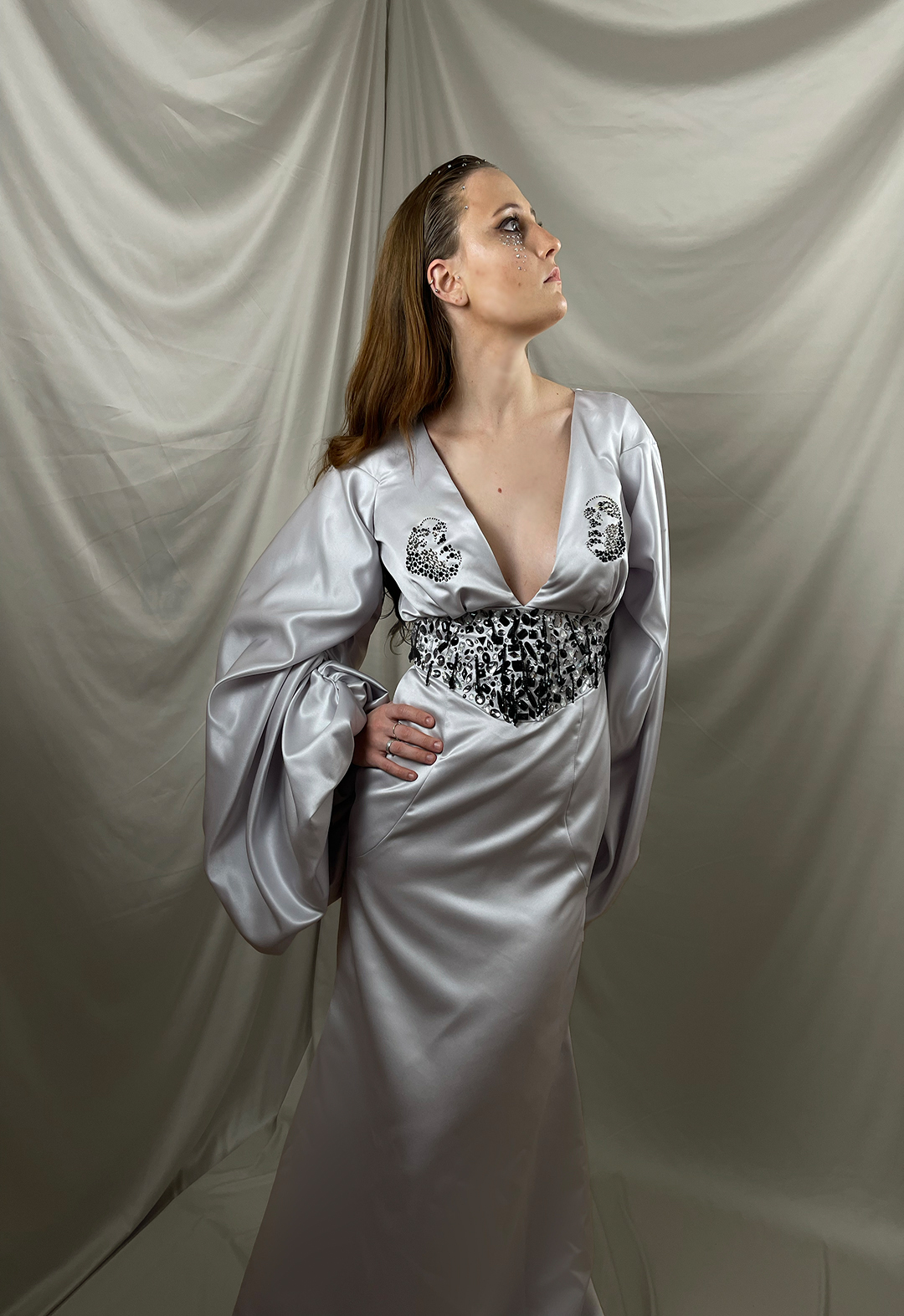 A model wearing a dress made in duchess satin, with rhinestones of different sizes and crystal of mixed shapes. The model has one hand on her waist and is looking to the side. The background has champagne colored silk drapes. 