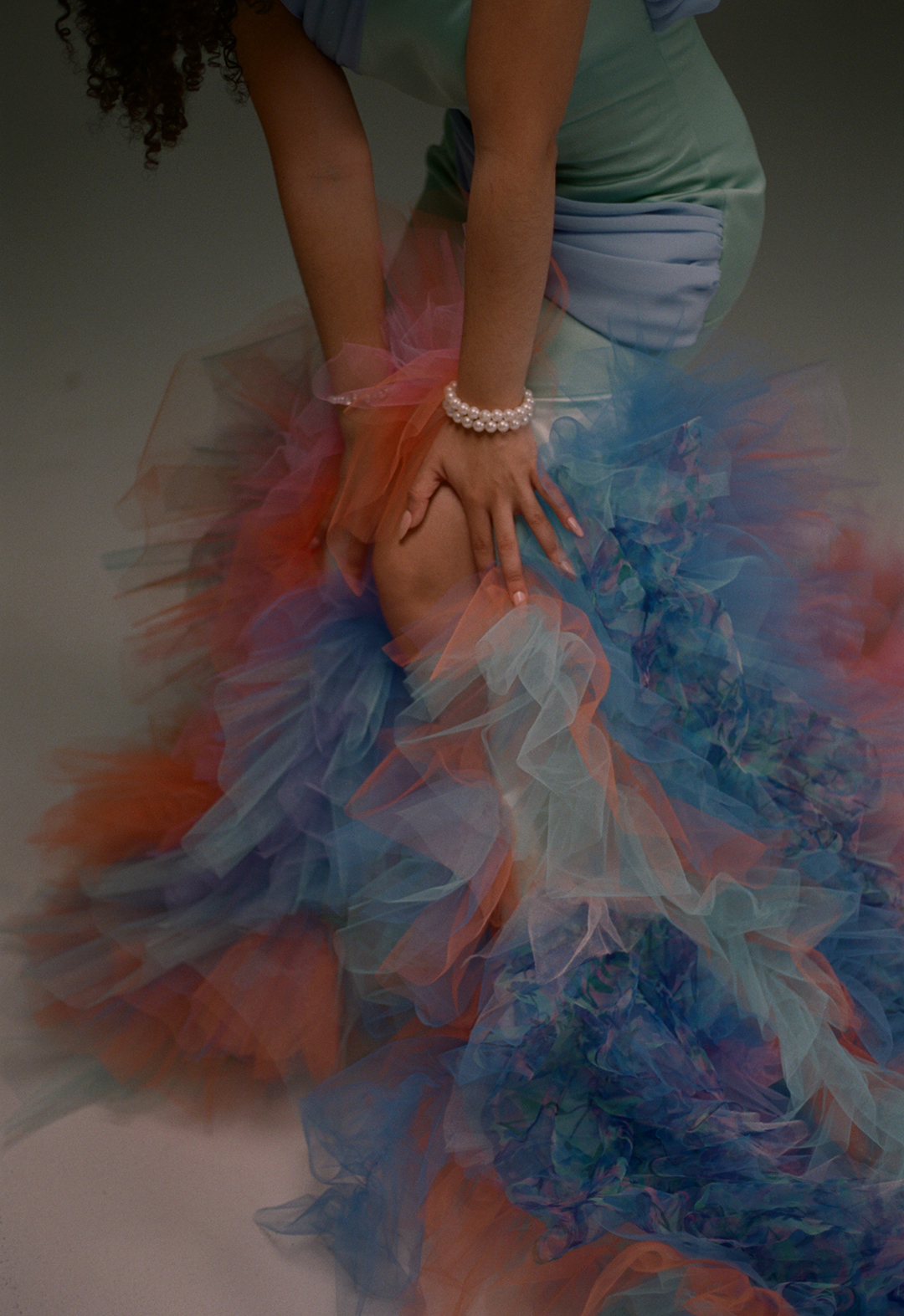 Zoomed in image of the bottom half of a tulle gown, where model is bending her knee to show the slit. Both her hands are on her knee. She is wearing two pearl bracelets on one wrist. 