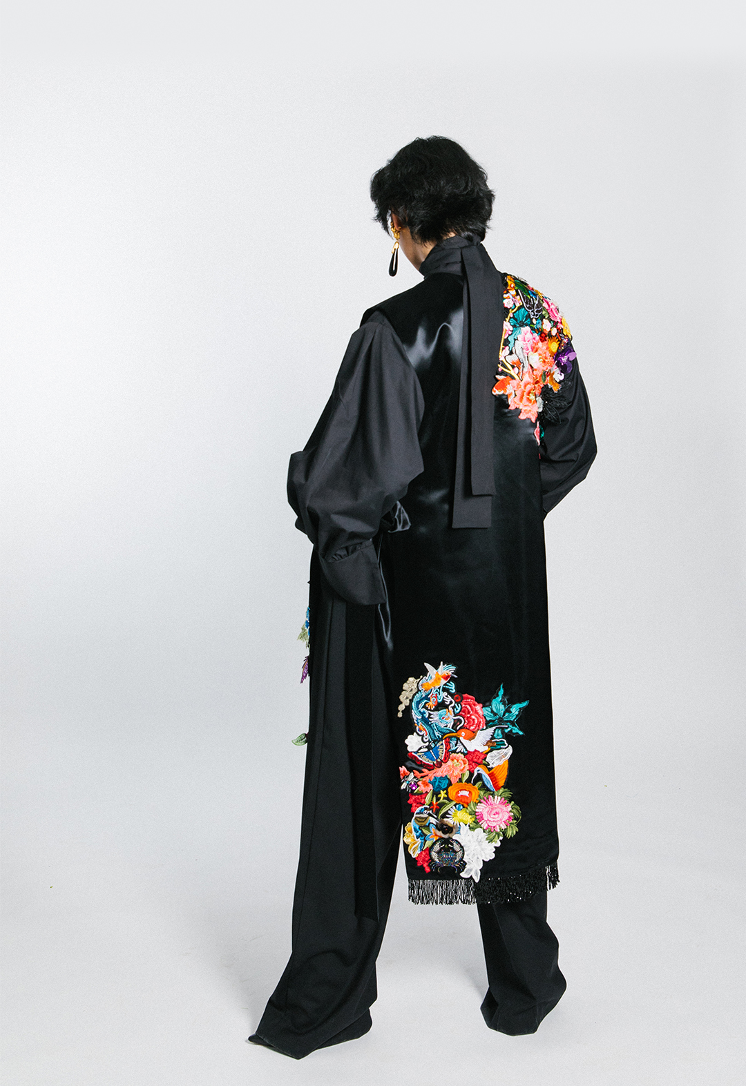 This is the back view of the model wearing black wide-legged wool pants with a black cotton poplin tie-neck blouse under a black satin poncho with collaged clusters and a printed lining.