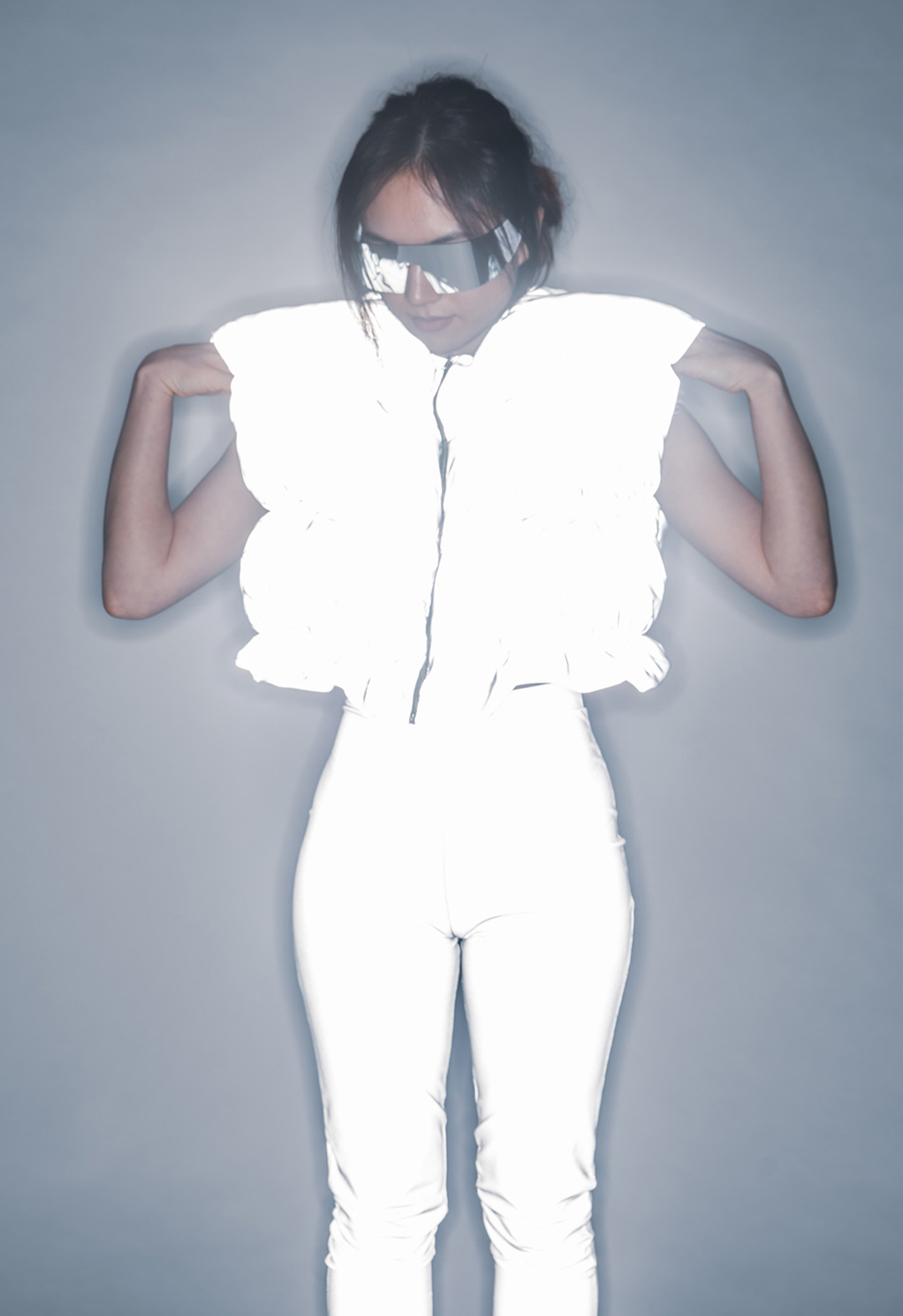 A model wearing a white vest and white pants, both made of fabric that is reflecting. The model is adjusting the shoulders of the vest. 