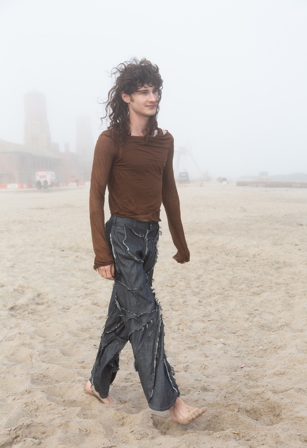 A model wears a two-way-stretch, soft-knit brown long-sleeved top, over a tank top in the same knit fabric, along with midweight denim pants of a blueish gray wash that have raw fraying edges, patched together by many pieces. 