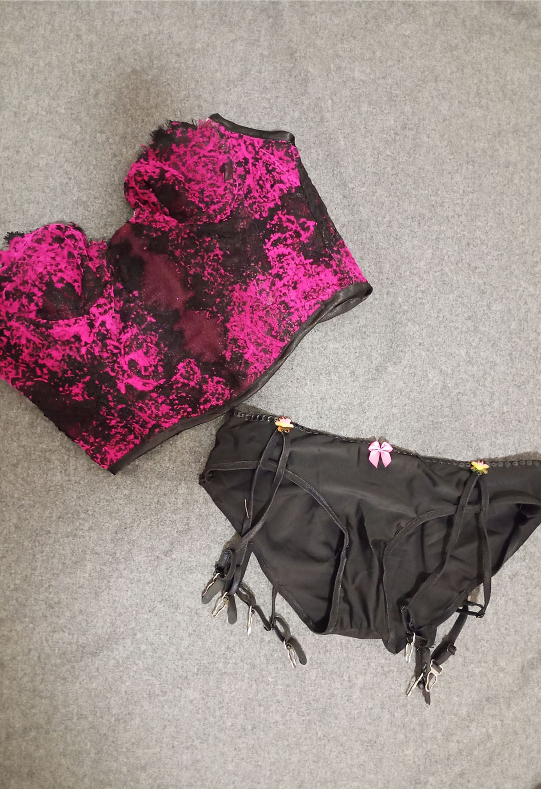 The photo shows a pink-and-black abstract lace bustier and a black panty with built-in garters lying on a gray fabric.