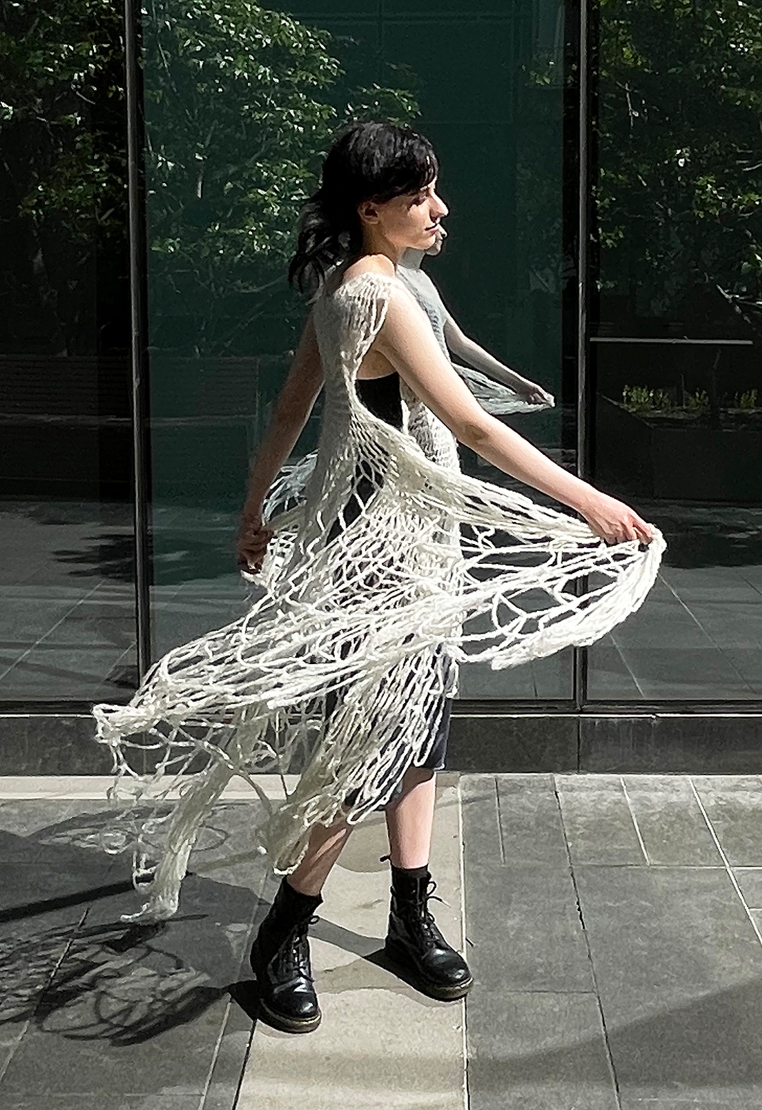 Side view of a model wearing a hand crocheted white lace suri alpaca dress. The model is swinging the hem of the dress. She is wearing combat boots and there is a glass building in the background, with reflection of trees.