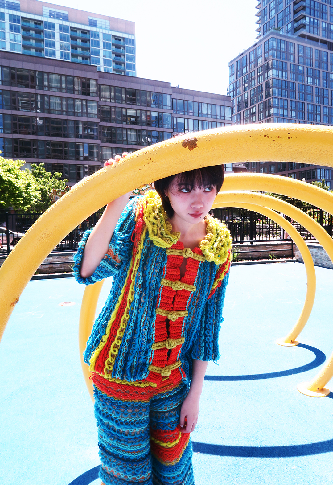 A model is standing in a playground, wearing a knit suit. The yarns alternate randomly between red, blue, and yellow. There is a river in the background.