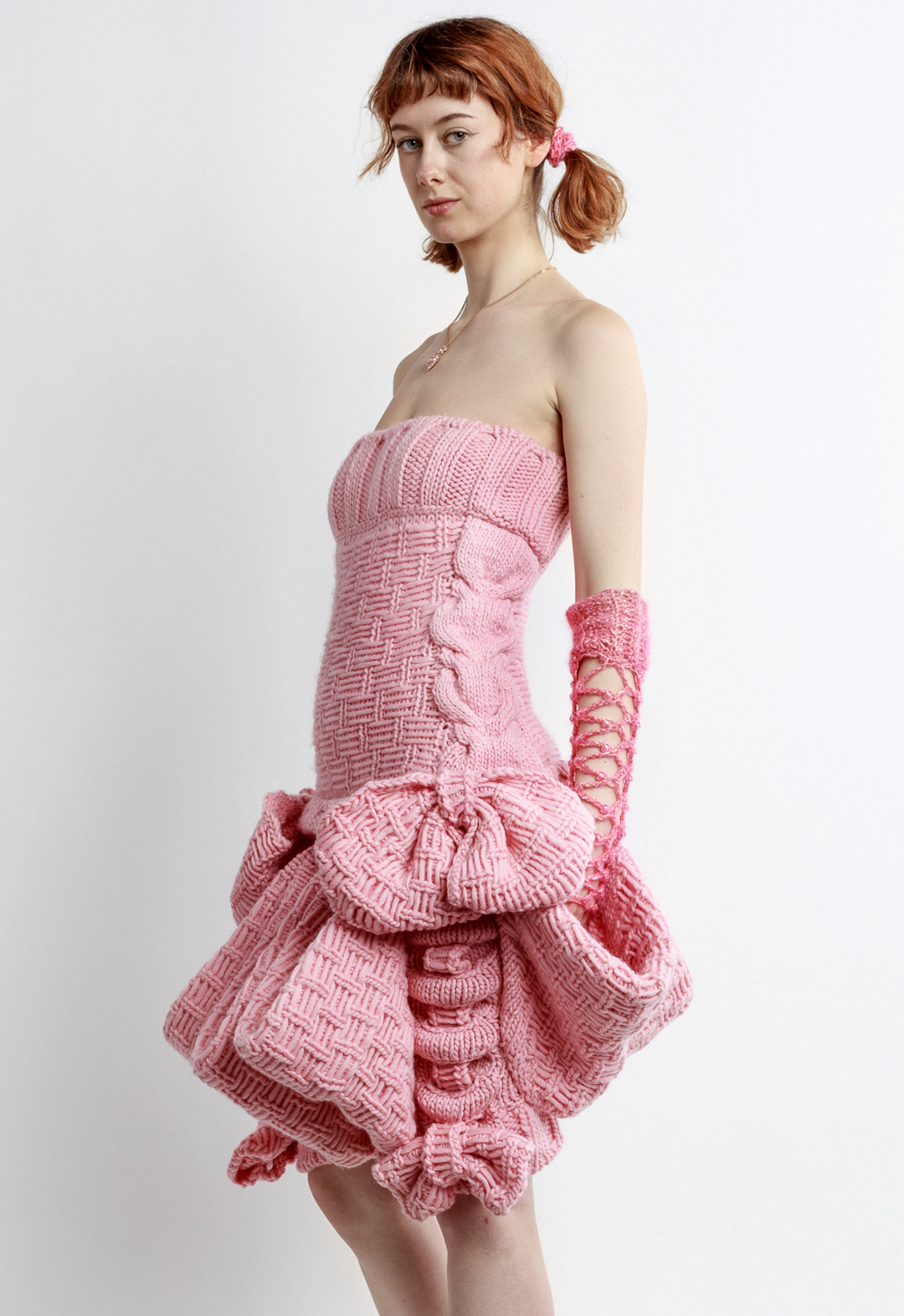 Photo of a girl posing in a pink sleeveless magical girl mini dress, looking to the camera. The dress is hand knit with baby pink pink wool. The dress features big bows and big pockets circling the skirt of the dress. The dress, and girl, are posed at a three-quarter view to the camera. The girl is wearing crochet fishnet gloves, pigtails, and a heart necklace. The girl has her hand placed in one of the big pockets.