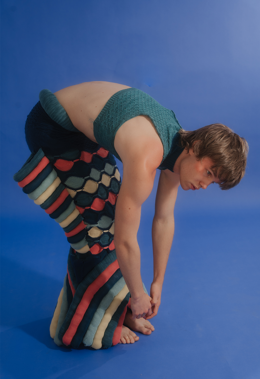 The photo shows a close-up shot of the model in a bent-over pose, wearing a sculptural knit skirt and tank top.