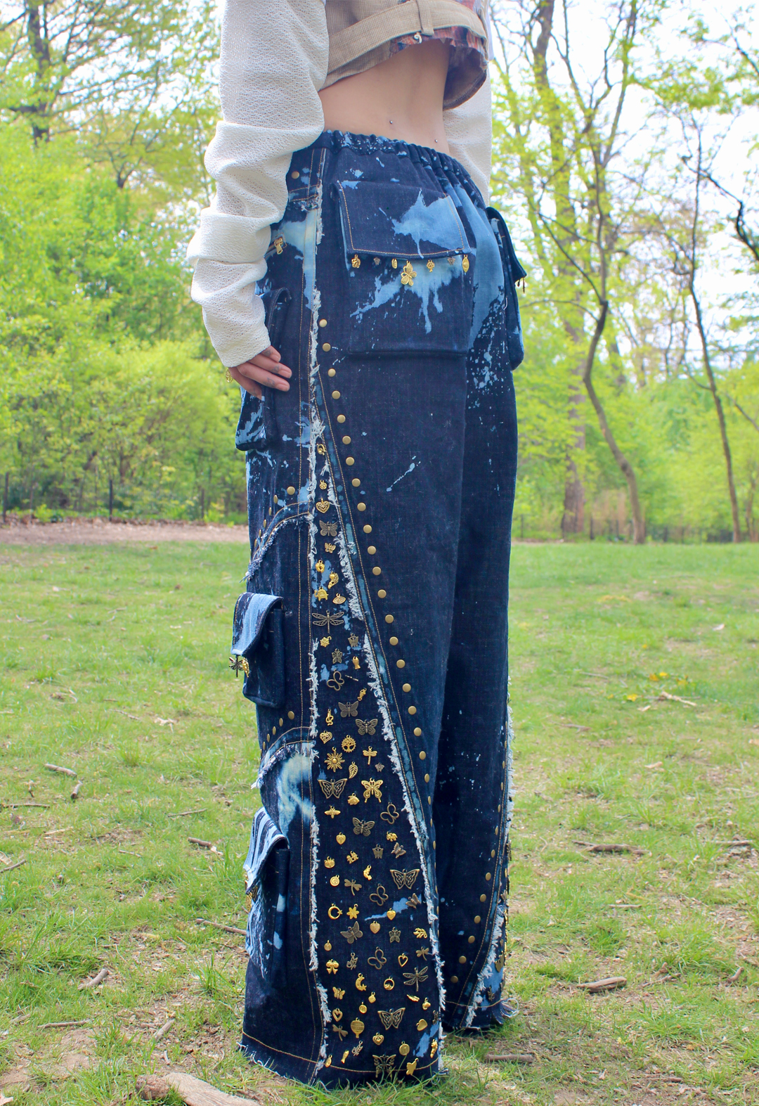 Three-quarter view of the baggy jean. Close-up view of  style lines with flat-fell seams and distressed edges. Bronze studding following along style lines. Hand-sewn gold and bronze charm embellishments along pocket flaps and filling side area. Charms include many shapes, such as butterflies, other insects, and flowers. 