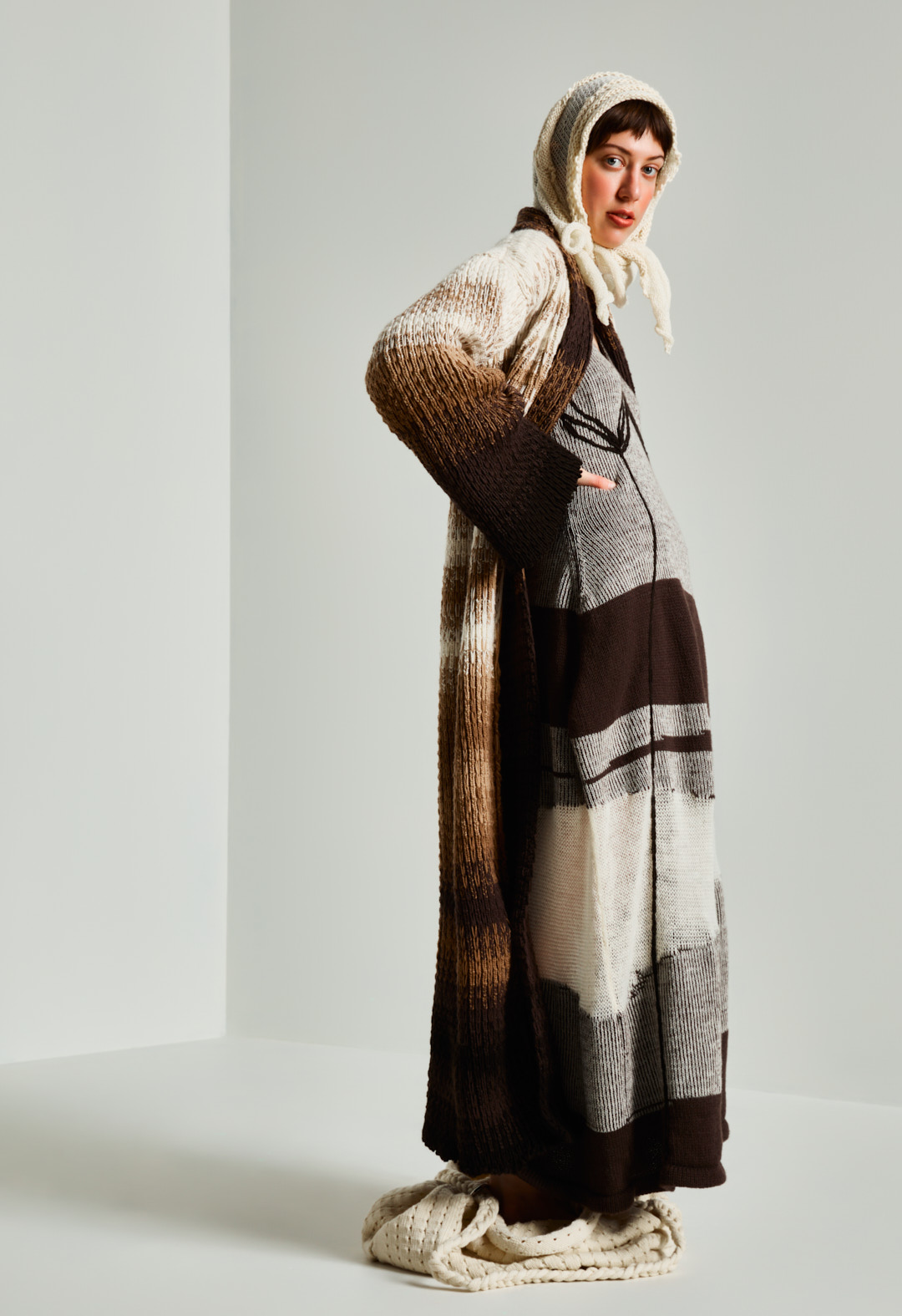 A full-length side-view shot of a model posing in a striped textured cardigan, made of organic certified merino over a midi dress, with a messenger bag and a handkerchief head covering. She is posing with her hand on her hip.