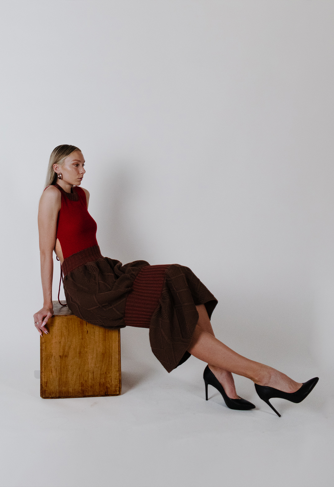 The same dress is displayed with a white background. This is a side-view photo of the model, who is sitting on a wooden box with her legs extended to the side. The skirt is beautifully draped across her legs. She is looking toward the side, with her face looking away from the camera and her hand propped behind her at the edge of the wooden box. 