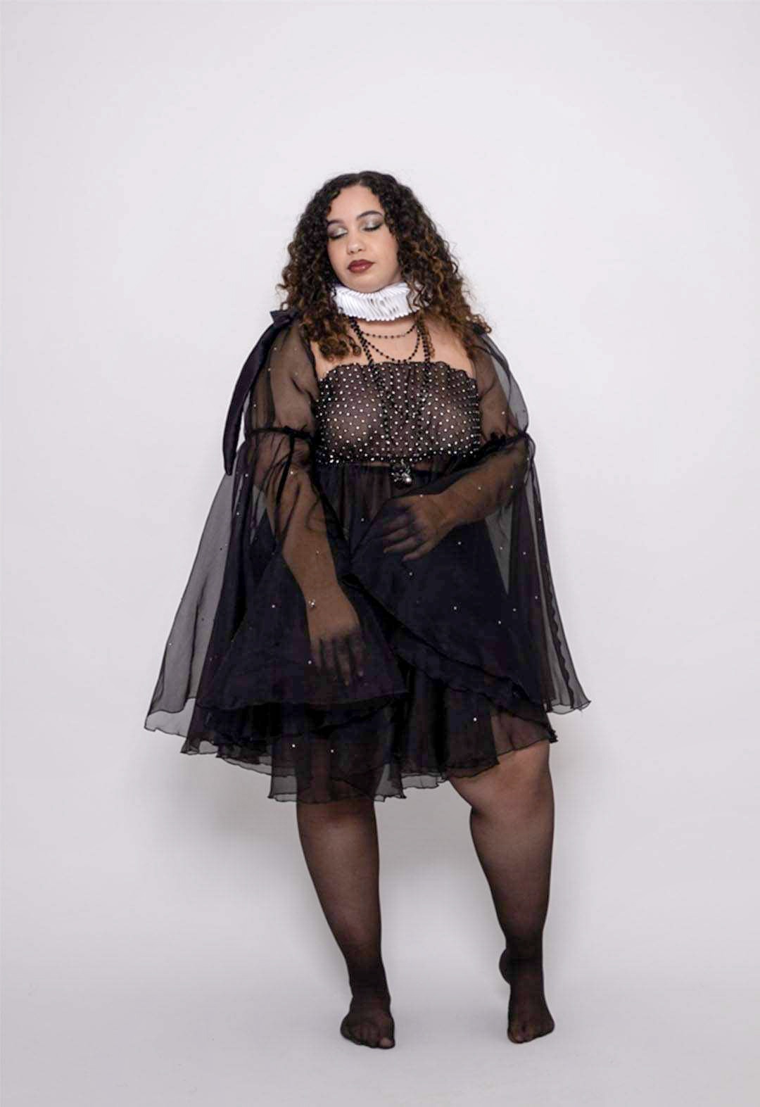 The model is wearing a black organza babydoll dress with rhinestone netting, over black mesh panty with chains, 