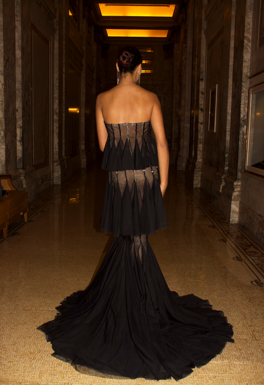 This is the back view of the black mesh three tiered gown. The gown has a train. In the image, the model is standing in a hallway with the main focus on the dress and a darker background. 