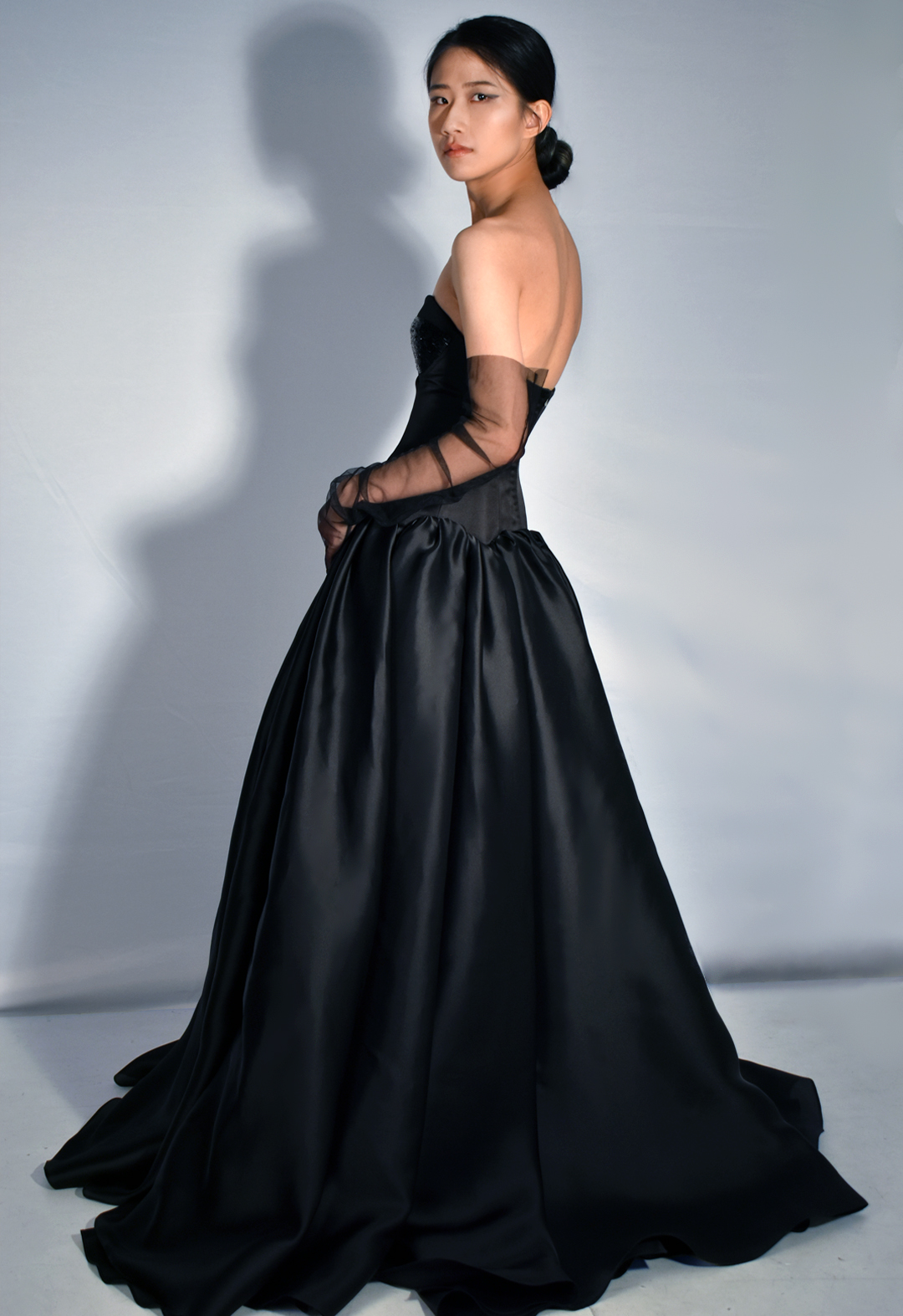 This black gown is made from 100 percent silk. The photo shows a three-quarter side view of a woman wearing a black silk gown with beading covering three-quarters of the bodice cups. The floor-length gown has a drop-waist duchesse satin bodice with cone-like satin-faced organza skirt panels gathered along the drop-waist, pooling onto the floor. The model is wearing black tulle gloves that cover three-quarters of her arms. Her face is facing the viewer. Her left arm rests along her hip, with her right hand in front of her. The background is white.