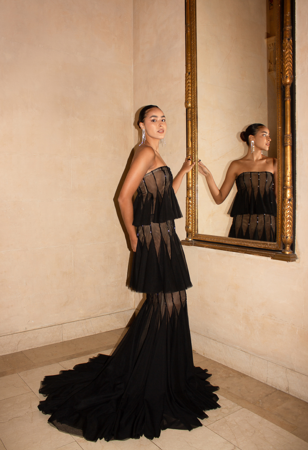 This tiered gown is made in black mesh fabric. In this image, the model is standing in front of a gold-framed mirror and looking into the camera. 