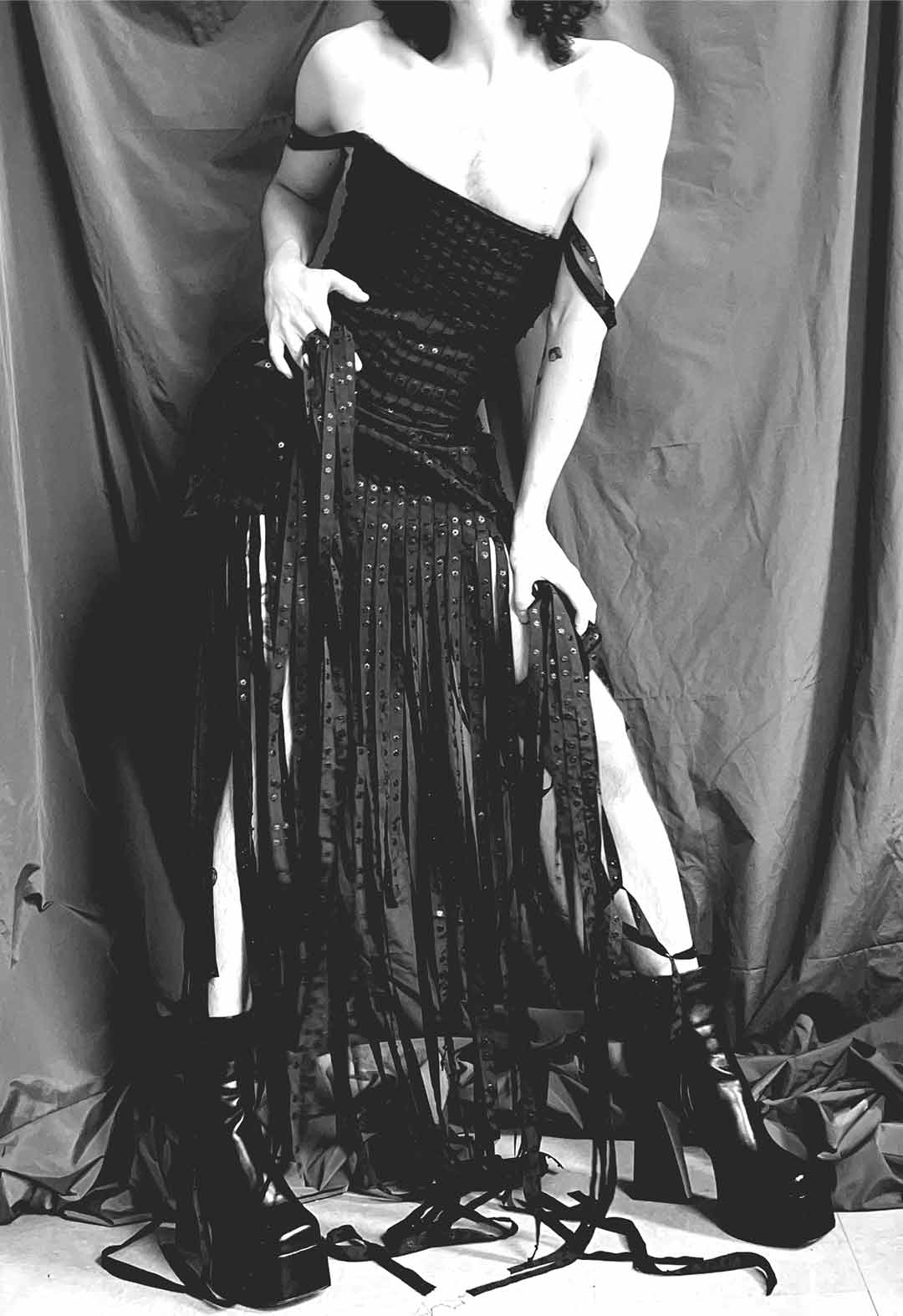 Detail view of model wearing black corset and matching miniskirt. The model is hunched over, and the body is a bit distorted. The model is holding some of the fringed pieces of the skirt.