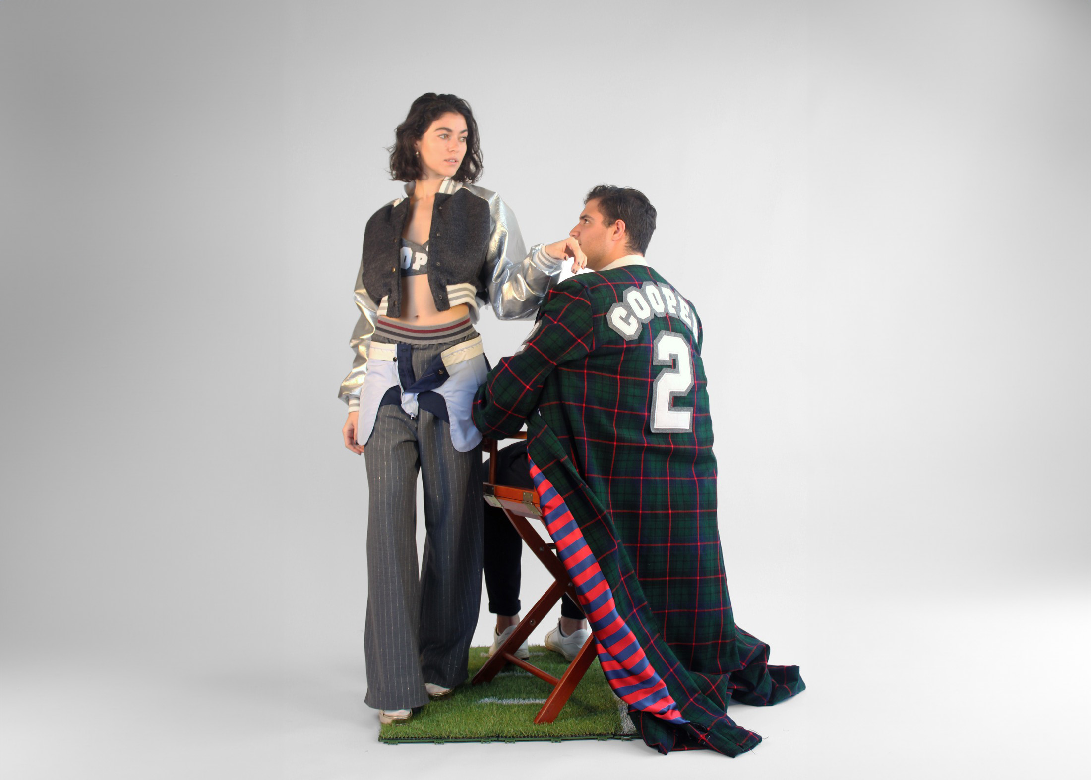 A male model in a plaid coat sits next to a female model wearing a silver cropped varsity jacket and pinstripe pants.
