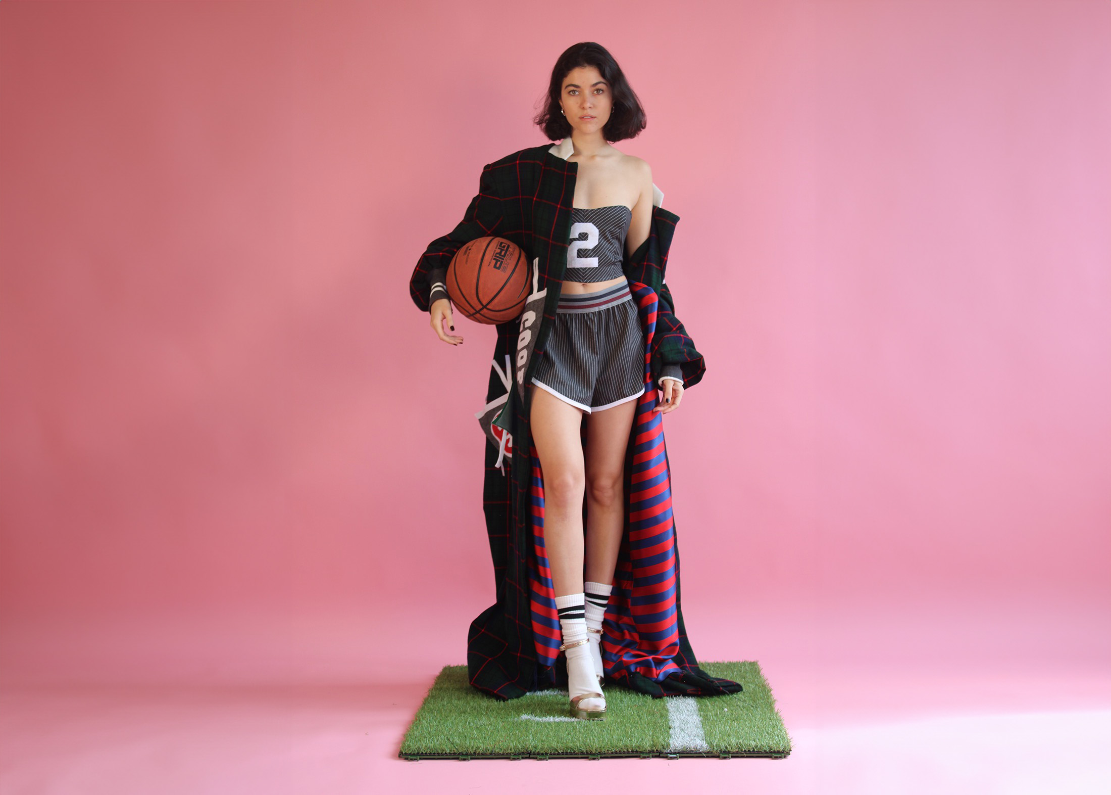 Caucasian model with dark hair models a green and red tartan coat with pennant appliqués. The floor length coat has a red and blue striped lining that shows through the model’s legs. She wears a sweetheart gray pinstripe bustier with a white number 2 on the front. The short matches the bustier and has a white trim on the hem.